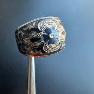May include: A silver ring with a black floral design. The ring is wide and has a textured surface.