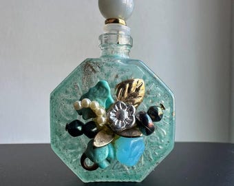 Könnte beinhalten: Eine Vintage-Glasflasche mit einem dekorativen Stopfen und einem Blumendesign aus Perlen und anderen Verzierungen. Die Flasche ist achteckig und hat eine mattierte, hellblaue Oberfläche.