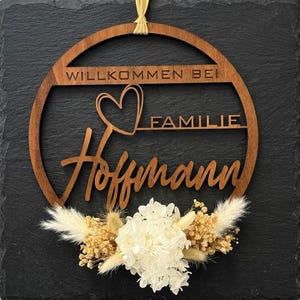 Könnte beinhalten: Ein kreisförmiges braunes Holzschild mit dem lasergeschnittenen Text WILLKOMMEN BEI FAMILIE Hoffmann und einer Herzform. Das Schild ist unten mit einem Arrangement aus getrockneten weißen Hortensien und beigen Pampasgraswedeln verziert.