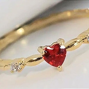 Peut inclure: Une bague en or avec une pierre rouge en forme de cœur et de petites pierres claires. La bague est délicate et a une bande torsadée.
