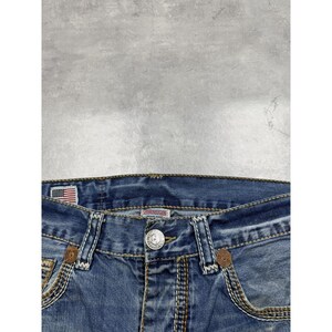 May include: Blue denim jeans featuring a silver metal button closure, thick tan contrast stitching, and a small American flag patch on the interior waistband. The jeans include two front pockets with brown circular accents and a central brand label.