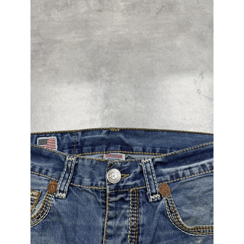 May include: Blue denim jeans featuring a silver metal button closure, thick tan contrast stitching, and a small American flag patch on the interior waistband. The jeans include two front pockets with brown circular accents and a central brand label.