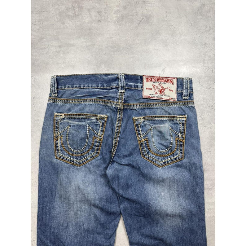 May include: Blue denim jeans featuring two back pockets with thick yellow and white stitching in a horseshoe pattern. A white rectangular patch with red text reading True Religion Brand Jeans World Tour is sewn above the right pocket.