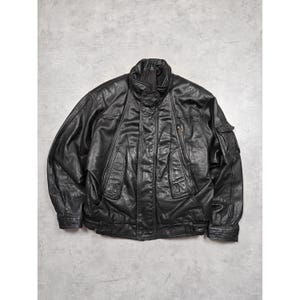 Könnte beinhalten: Schwarze Lederjacke mit hohem Kragen, mehreren Taschen und Knopfleiste. Die Jacke hat ein klassisches Design mit leicht gepufften Ärmeln und geripptem Saum. Das Leder scheint weich und abgenutzt zu sein.