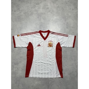May include: White and red Spain soccer jersey with a V-neck collar and short sleeves. The jersey features the Adidas logo, the Spanish coat of arms, and the word "ESPAÑA". Red accents are on the sleeves and sides.