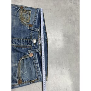 May include: Blue denim jeans featuring a faded wash, contrast stitching, and copper-toned metal rivets. A white measuring tape is placed vertically along the side, showing a length measurement extending past 45 centimeters on the fabric.