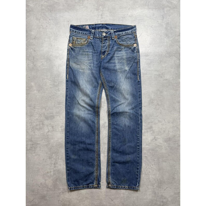 May include: Blue denim jeans featuring a faded wash, a button fly closure, and prominent gold-tone contrast stitching along the seams and pockets. The jeans include five pockets, belt loops, and a metal button at the waist with a classic straight leg cut.