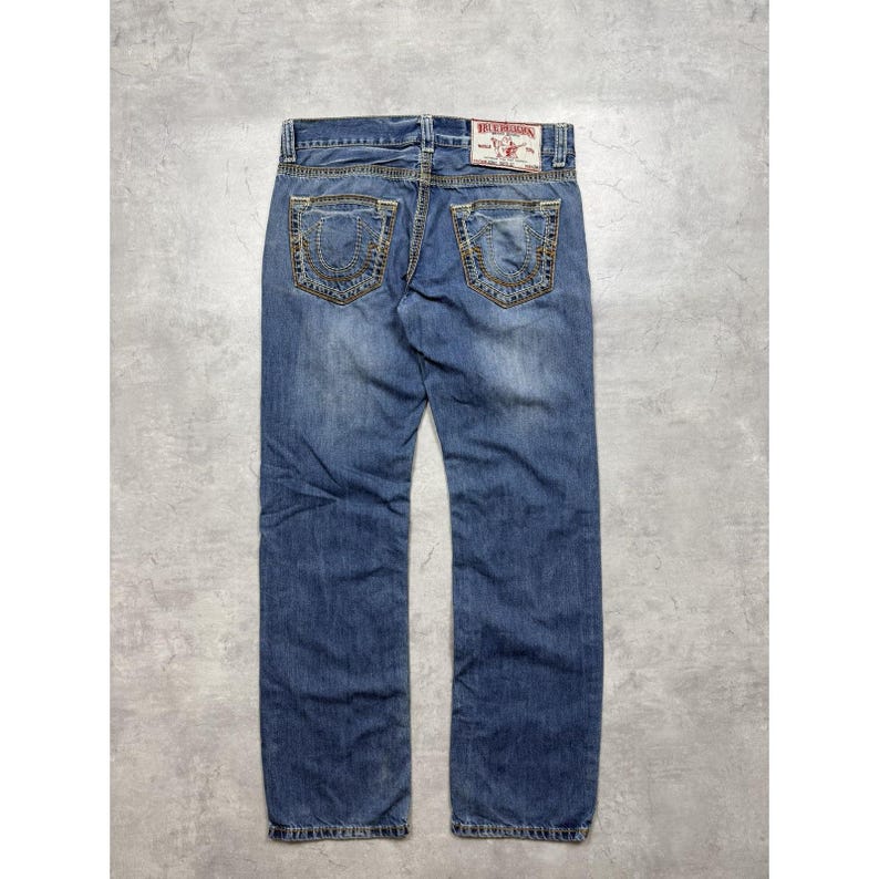 May include: Blue denim jeans featuring a medium wash, a button fly, and two back pockets with decorative gold-tone stitching. A white rectangular brand label with red text is attached to the back waistband above the right pocket.