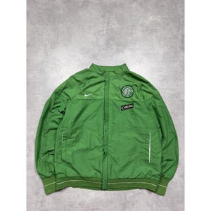 Könnte beinhalten: Grüne Sportjacke mit weißem Nike-Logo und Vereinswappen auf der Brust. Die Jacke hat einen gerippten Kragen und Bündchen mit weißen Streifen. Das Wort "Carling" ist auf einem Aufnäher aufgedruckt.