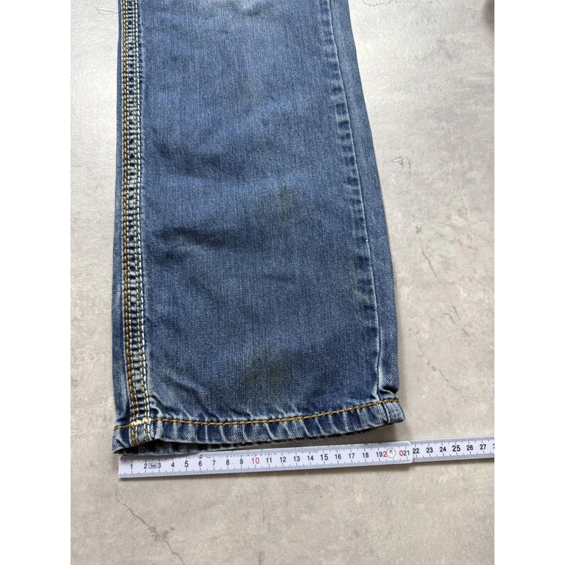 May include: A blue denim pant leg featuring a vertical side seam with decorative gold and white stitching. A white measuring tape is positioned horizontally at the hem, showing a width measurement of approximately 8.25 inches across the fabric.