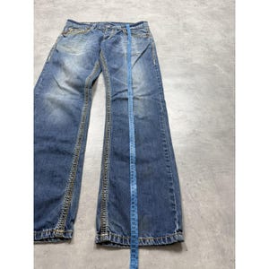 May include: Blue denim jeans featuring a faded wash, contrast stitching, and a button fly. A blue measuring tape is placed vertically along the center of the leg, showing measurements up to 112 centimeters, which is approximately 44.09 inches.