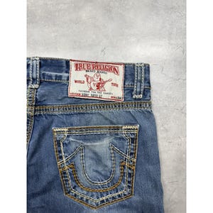 May include: Blue denim jeans featuring thick, contrasting gold-tone stitching on the back pocket and waistband. A white rectangular patch is sewn on the back, displaying red text that reads True Religion Brand Jeans World Tour Bobby Super QT.