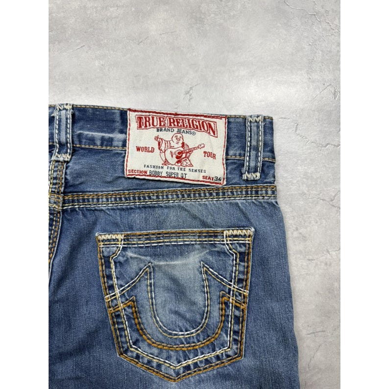 May include: Blue denim jeans featuring thick, contrasting gold-tone stitching on the back pocket and waistband. A white rectangular patch is sewn on the back, displaying red text that reads True Religion Brand Jeans World Tour Bobby Super QT.