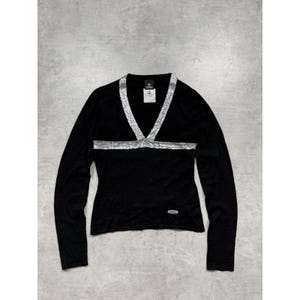 Puede incluir: Top negro de manga larga con cuello en V y ribete de lentejuelas plateadas en el escote y el pecho. El top tiene una silueta ajustada y un pequeño logotipo cerca del dobladillo. Una etiqueta es visible cerca del cuello.