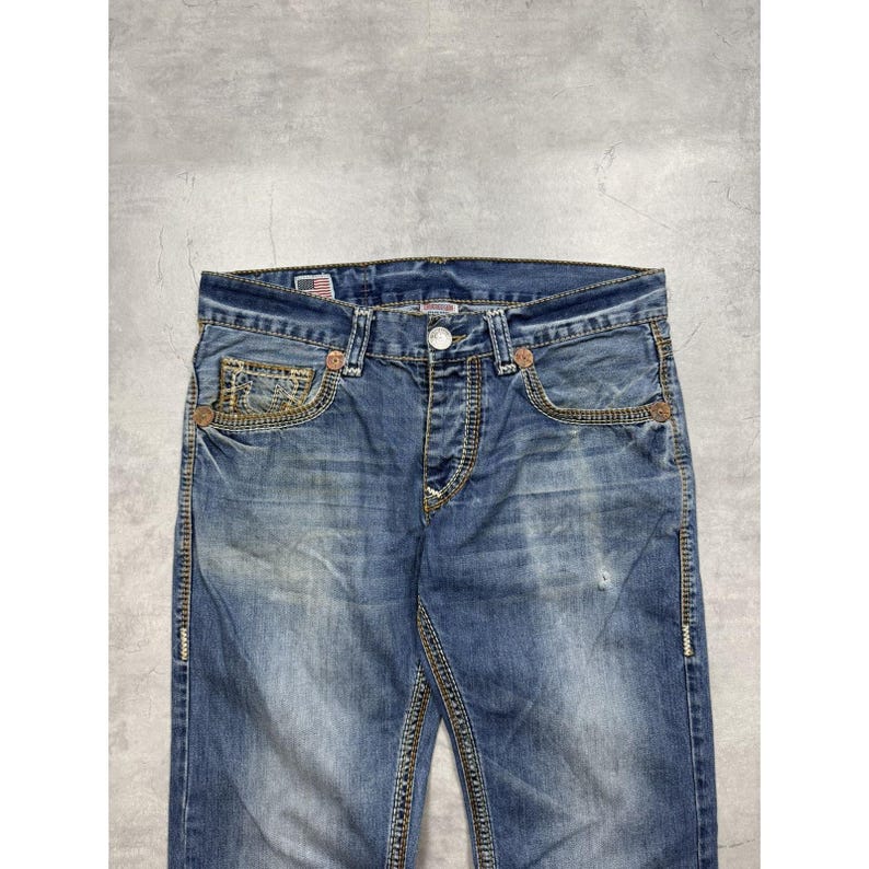 May include: Blue denim jeans featuring a faded wash, copper-toned rivets, and thick contrast stitching. The waistband includes a small American flag patch near the front pocket, a button closure, and a zipper fly with five-pocket styling.