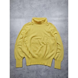 Può includere: Un maglione giallo senape a collo alto con maniche lunghe. Il maglione è realizzato in morbido materiale a maglia e ha orlo e polsini a coste. Il collo alto è ripiegato. Il maglione è esposto su una superficie grigia.