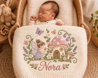 May include: A cream-colored knit baby blanket featuring an embroidered scene of a fairy, a mushroom house, flowers, and butterflies. The name Nora is embroidered in pink script at the bottom. The blanket is draped over a baby lying in a bassinet.