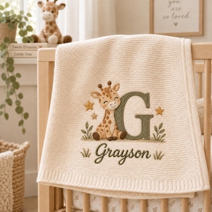 Peut inclure: Une couverture pour bébé en tricot couleur crème avec une broderie représentant une girafe brune assise à côté d'une grande lettre G vert olive. Le prénom Grayson est brodé en lettres cursives vert olive sous la girafe, avec des étoiles.