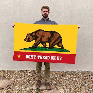 May include: A rectangular flag featuring a brown bear walking on green grass against a yellow background. The bottom section is red with a gold star and the text "DON'T TREAD ON US".