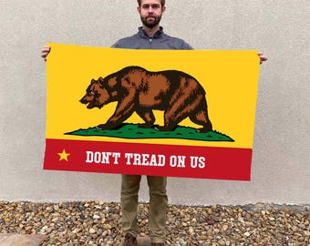 May include: A rectangular flag featuring a brown bear walking on green grass against a yellow background. The bottom section is red with a gold star and the text "DON'T TREAD ON US".