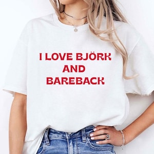 Puede incluir: Camiseta blanca con el texto rojo "I LOVE BJÖRK AND BAREBACK". La camiseta tiene cuello redondo y mangas cortas. El texto está centrado en la parte delantera de la camiseta.