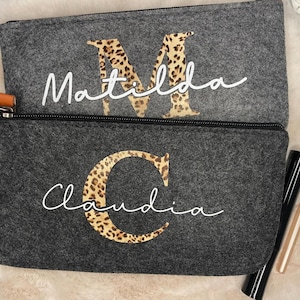 Könnte beinhalten: Zwei graue Kosmetiktaschen aus Filz mit schwarzen Reißverschlüssen. Eine Tasche hat den Namen "Matilda" mit einem Leopardenmuster "M". Die andere Tasche hat den Namen "Claudia" mit einem Leopardenmuster "C". Make-up-Artikel liegen neben den Taschen.