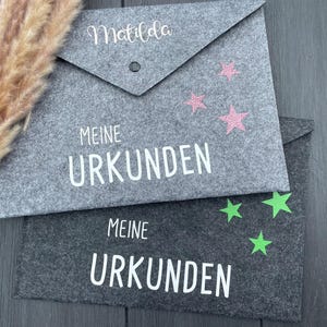 Könnte beinhalten: Zwei graue Filz-Dokumentenhalter. Der obere hat den Namen "Matilda" und rosa Glitzersterne. Der untere hat grüne Sterne. Beide haben die Worte "MEINE URKUNDEN" in Weiß.
