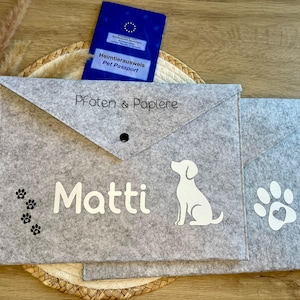 Könnte beinhalten: Zwei hellgraue Dokumentenmappen aus Filz mit dreieckiger Klappe und schwarzem Druckknopfverschluss. Die vordere Mappe zeigt den Text "Pfoten & Papiere", den Namen "Matti", eine weiße Hundesilhouette und kleine schwarze Pfotenabdrücke.