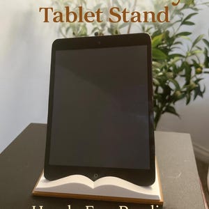 May include: A black tablet is displayed on a white and brown book-shaped stand. The stand is on a dark surface. Text on the image reads "E-Book Buddy Tablet Stand" and "Hands-Free Reading, Anywhere."