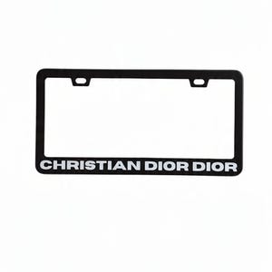 May include: Black license plate frame with the text "CHRISTIAN DIOR DIOR" in white. The frame has a rectangular shape with two mounting holes at the top. The background is plain white.