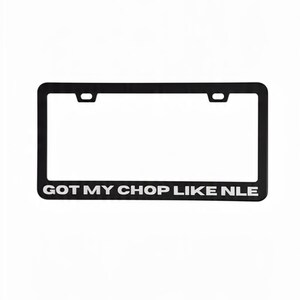 May include: Black license plate frame with the text "GOT MY CHOP LIKE NLE" in white. The frame has a rectangular shape with two mounting holes at the top. The text is centered at the bottom of the frame.