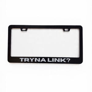 Puede incluir: Marco negro para matrícula con el texto blanco "TRYNA LINK?". El marco tiene forma rectangular con esquinas redondeadas y dos orificios de montaje en la parte superior. El fondo es blanco.