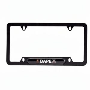 May include: Black license plate frame with the word "BAPE" in white lettering. The frame has a rectangular shape with rounded corners and mounting holes. The BAPE logo is also visible.