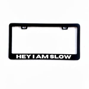 Puede incluir: Marco negro para matrícula con el texto "HEY I AM SLOW" en blanco. El marco tiene forma rectangular con esquinas redondeadas y dos orificios de montaje en la parte superior.