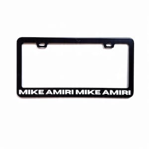 May include: Black license plate frame with the text "MIKE AMIRI" in white. The frame is rectangular with rounded corners and two mounting holes at the top. The text is centered at the bottom.
