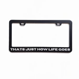 May include: Black license plate frame with the text "THATS JUST HOW LIFE GOES" in white lettering. The frame has a rectangular shape with two mounting holes at the top. The background is plain white.