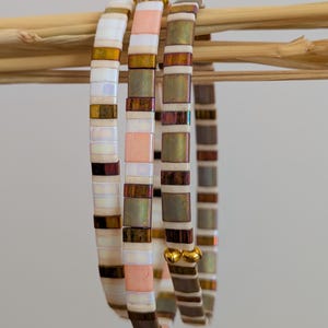 Peut inclure: Trois bracelets extensibles composés de petites perles plates et carrées. Les perles présentent un mélange de finitions blanc irisé, pêche mat et bronze métallique. Chaque bracelet est terminé par deux petites perles métalliques dorées.