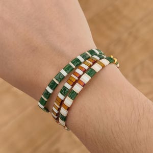 Peut inclure: Trois bracelets empilés avec des carreaux verts, blancs et orange. Les bracelets sont au poignet d'une personne.