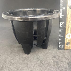 May include: A black plastic plant pot with a raised lip around the top and a series of vertical slits around the sides. The pot has a rounded bottom with four small feet.