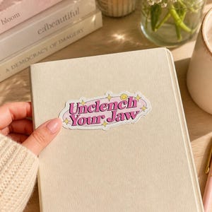 May include: A rectangular sticker with rounded edges featuring the text Unclench Your Jaw in bold pink letters. The design includes a light pink oval background, small yellow four-pointed stars, and a tiny yellow smiley face icon on a white notebook.