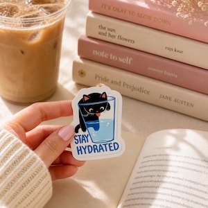 May include: A die-cut vinyl sticker featuring a black cat with its head and paws inside a clear glass of water, with its tongue sticking out. The text "STAY HYDRATED" is printed in blue capital letters at the bottom of the sticker design.