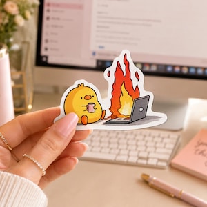 May include: A die-cut vinyl sticker featuring a yellow cartoon chick sitting on the ground holding a pink mug. Next to the chick is a black laptop engulfed in bright orange and red flames. The sticker has a white border and a glossy finish.