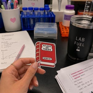 以下が含まれることがあります： ピンクのしおりが付いた赤い本型の長方形ステッカー。表紙には黒い文字で「BUT WOULD YOUR THESIS DEFEND YOU?」と書かれています。ステッカーには白い縁取りがあり、実験器具を背景にして持たれています。