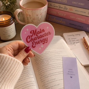 May include: A heart-shaped sticker with a pink background and white sparkles featuring the red text Main Character Energy. The sticker is held over an open book next to a lavender bookmark with the text one more chapter and a stack of books.