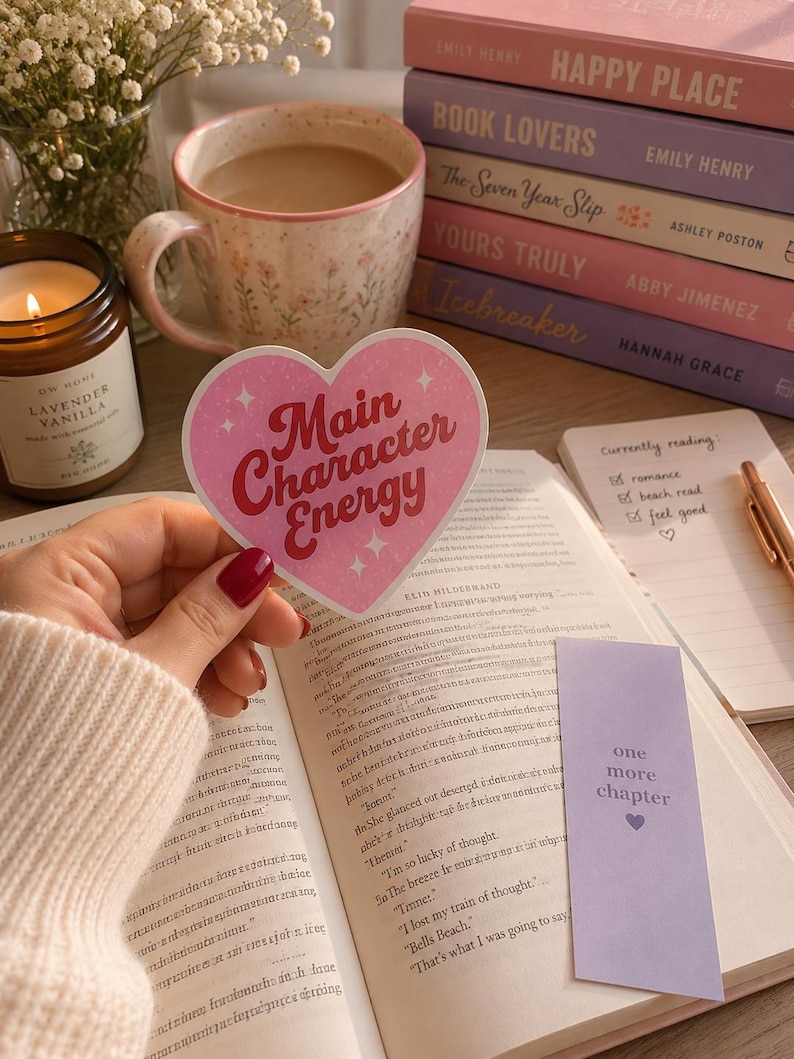 May include: A heart-shaped sticker with a pink background and white sparkles featuring the red text Main Character Energy. The sticker is held over an open book next to a lavender bookmark with the text one more chapter and a stack of books.