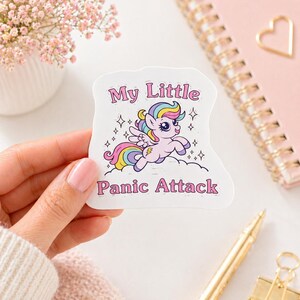 Puede incluir: Una pegatina troquelada que muestra un poni alado de color morado claro con crines y cola de colores arcoíris, volando sobre una nube. La pegatina incluye el texto "My Little Panic Attack" en letras rosas, rodeado de pequeñas estrellas negras.