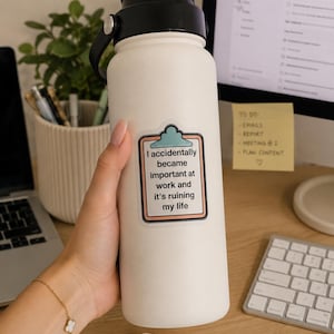 Può includere: Una borraccia termica bianca con tappo nero e un adesivo raffigurante un blocco per appunti. Il testo recita: I accidentally became important at work and it's ruining my life. La borraccia presenta una finitura opaca liscia e un manico.
