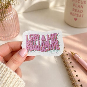 May include: A die-cut sticker with a white background featuring the text I CRY A LOT BUT I AM SO PRODUCTIVE IT'S AN ART in a retro, multi-layered font with pink, purple, and teal colors, accented by small star shapes around the lettering.