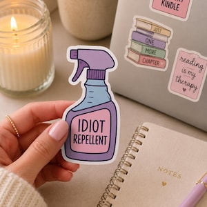 May include: A die-cut vinyl sticker shaped like a spray bottle with a purple nozzle and base, a light blue middle section, and a pink label featuring the text IDIOT REPELLENT in bold black capital letters. The sticker has a clean white border.