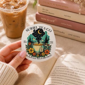 May include: A die-cut sticker featuring a tea cup with steam rising into a crescent moon and stars. The illustration includes mountains, pine trees, and colorful flowers. Text reads: SURIEL TEA CO, SPILL THE TEA, and THE HOTTEST TEA IN PRYTHIAN.